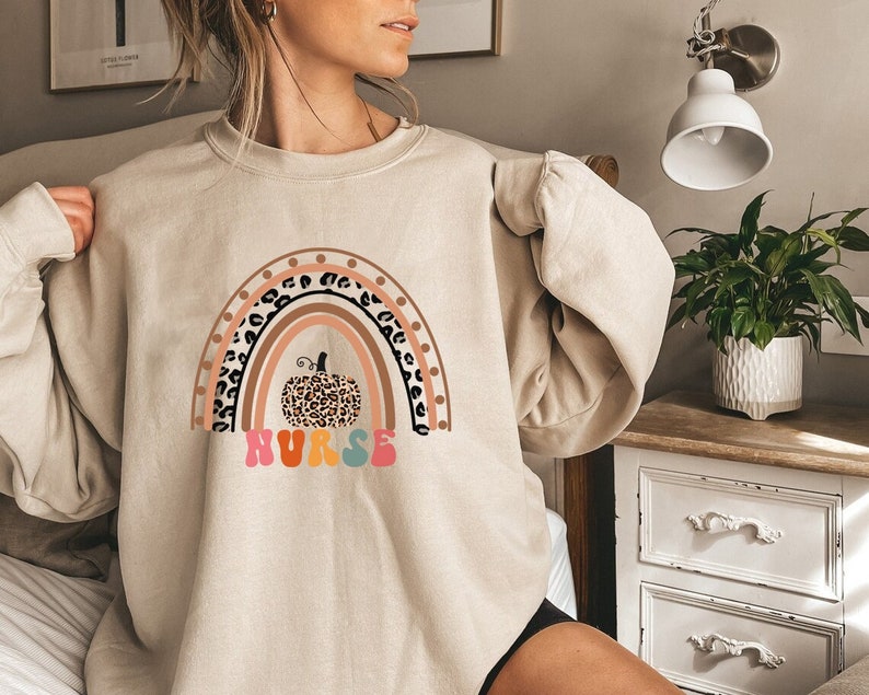 May include: A beige sweatshirt with a rainbow and a leopard print pumpkin. The rainbow has brown, pink, orange, yellow, and blue stripes. The text "NURSE" is written in rainbow colors below the pumpkin.