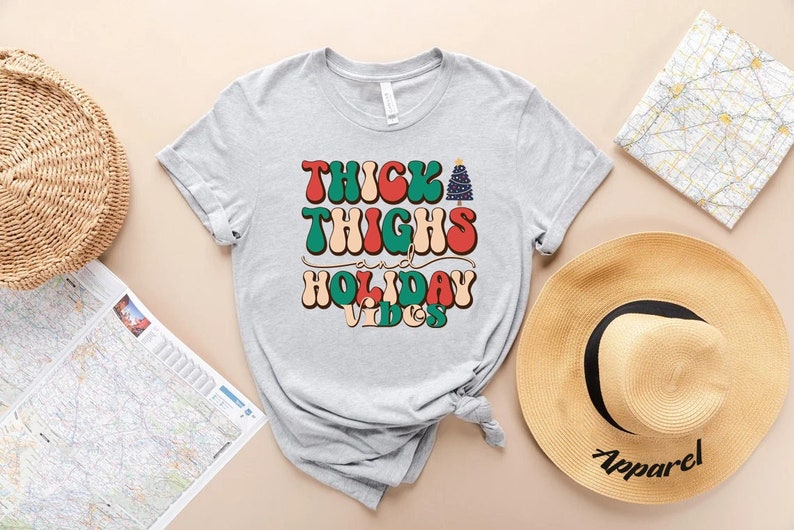 Puede incluir: Una camiseta gris con el texto "Thick Thighs and Holiday Vibes" en una fuente retro con un gr&aacute;fico de &aacute;rbol de Navidad.