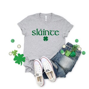 May include: A grey t-shirt with the Irish word "sl&aacute;inte" printed in green, along with a green four-leaf clover. The shirt is paired with blue jeans, white trainers, and green shamrock-shaped accessories.