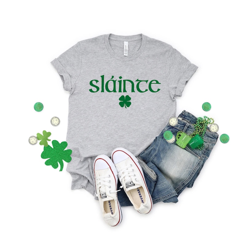 May include: A grey t-shirt with the Irish word "sl&aacute;inte" printed in green, along with a green four-leaf clover. The shirt is paired with blue jeans, white trainers, and green shamrock-shaped accessories.