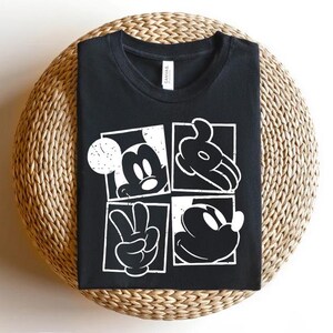 May include: Black t-shirt featuring a white graphic of Mickey Mouse in four square panels. The panels show Mickey's face, hand, and a peace sign. The shirt is displayed on a woven, circular surface.