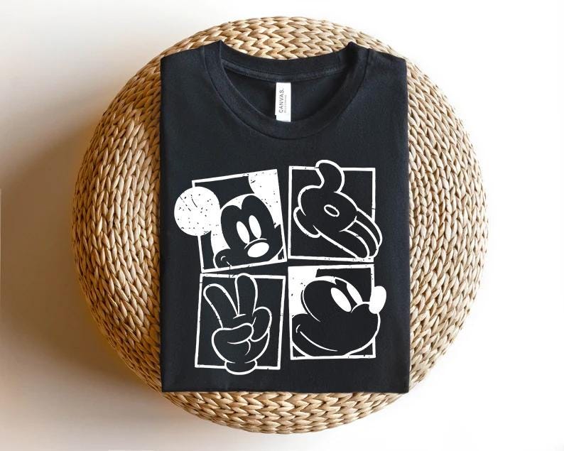 May include: Black t-shirt featuring a white graphic of Mickey Mouse in four square panels. The panels show Mickey's face, hand, and a peace sign. The shirt is displayed on a woven, circular surface.
