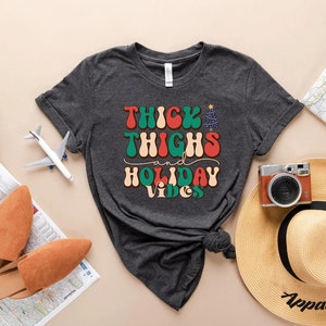 Puede incluir: Una camiseta gris oscuro con el texto "Thick Thighs and Holiday Vibes" en una fuente retro. El texto est&aacute; en rojo, verde y blanco. La camiseta est&aacute; anudada en la parte inferior.