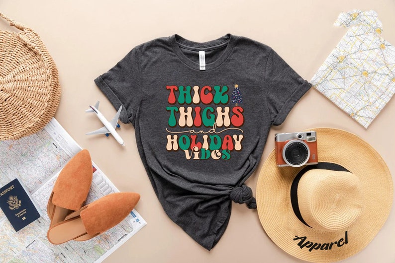 Puede incluir: Una camiseta gris oscuro con el texto "Thick Thighs and Holiday Vibes" en una fuente retro. El texto est&aacute; en rojo, verde y blanco. La camiseta est&aacute; anudada en la parte inferior.