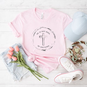 Op de afbeelding: Een lichtroze t-shirt met een cirkelvormige afbeelding van een kruis en de tekst "HE IS NOT HERE, HE HAS RISEN." De afbeelding bevat ook een lichtblauwe spijkerbroek, een lichtblauwe baseballpet, witte sneakers en een krans.