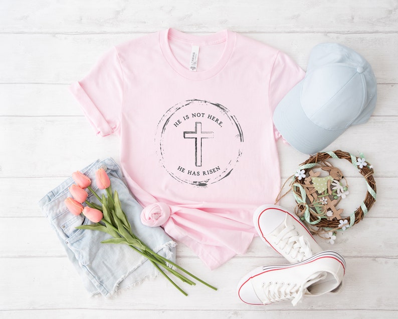 Op de afbeelding: Een lichtroze t-shirt met een cirkelvormige afbeelding van een kruis en de tekst "HE IS NOT HERE, HE HAS RISEN." De afbeelding bevat ook een lichtblauwe spijkerbroek, een lichtblauwe baseballpet, witte sneakers en een krans.