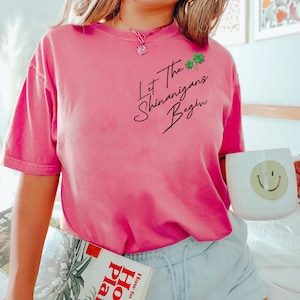 Peut inclure: Un t-shirt rose avec le texte "Let the Shenanigans Begin" en &eacute;criture noire avec un graphique de tr&egrave;fle vert. Le mod&egrave;le porte le t-shirt avec un short bleu et tient un livre intitul&eacute; "How to Grow House Plants".