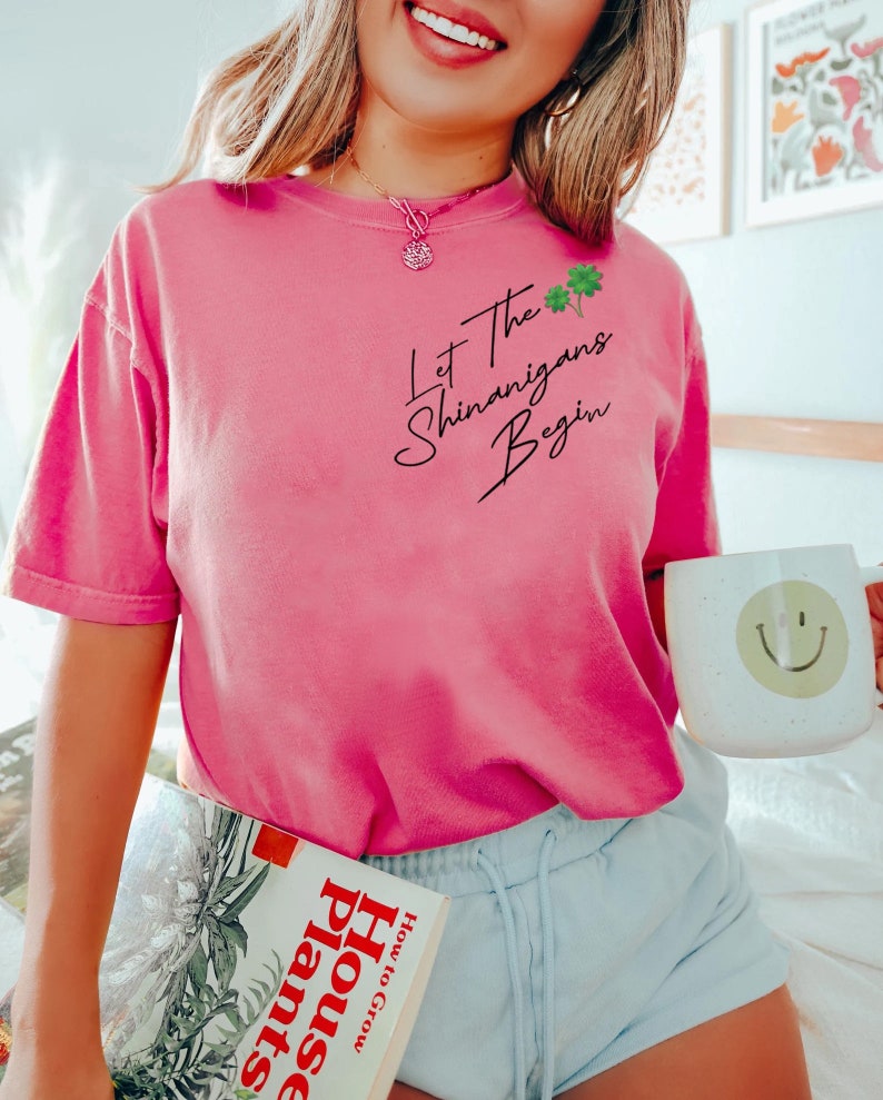 Peut inclure: Un t-shirt rose avec le texte "Let the Shenanigans Begin" en &eacute;criture noire avec un graphique de tr&egrave;fle vert. Le mod&egrave;le porte le t-shirt avec un short bleu et tient un livre intitul&eacute; "How to Grow House Plants".