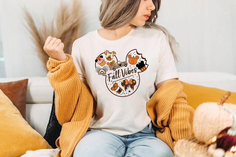 May include: A cream-coloured t-shirt with a Mickey Mouse head design, incorporating autumn-themed graphics. The design includes pumpkins, cupcakes, and the text "Fall Vibes". The person is wearing a mustard-coloured sweater.