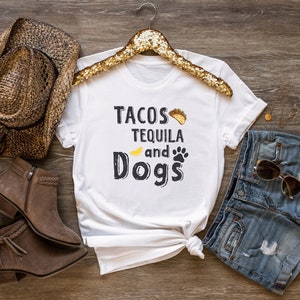 May include: White t-shirt with the text "Tacos Tequila and Dogs" in black lettering. The shirt is styled with a brown cowboy hat, brown boots, blue denim shorts, and gold sunglasses.