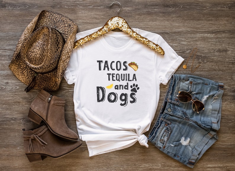 May include: White t-shirt with the text "Tacos Tequila and Dogs" in black lettering. The shirt is styled with a brown cowboy hat, brown boots, blue denim shorts, and gold sunglasses.
