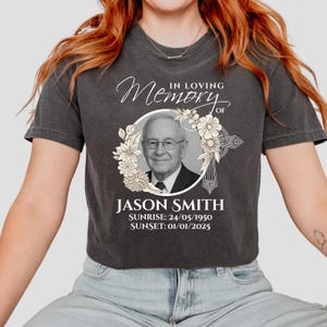 May include: Dark grey t-shirt with a memorial design. The design includes a portrait of a man within a floral wreath, the text "In Loving Memory of Jason Smith," and dates of birth and passing. The shirt is a casual wear item.