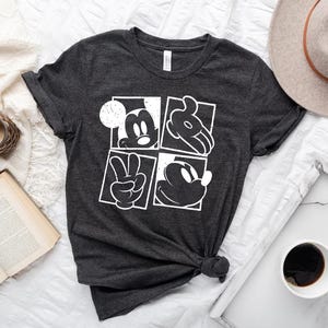 May include: Dark gray t-shirt featuring a white graphic of Mickey Mouse in four squares. The design includes Mickey's face, hands, and silhouette. The shirt is knotted at the bottom and is displayed on a white surface with a hat, coffee, and a book.