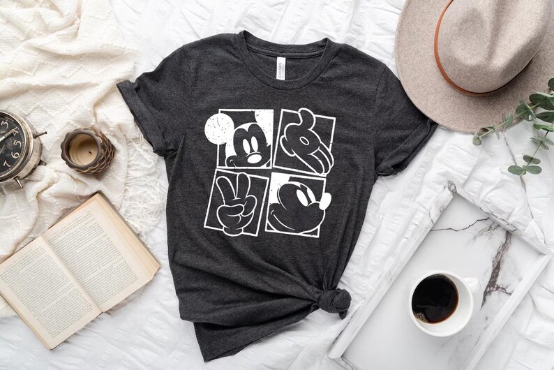 May include: Dark gray t-shirt featuring a white graphic of Mickey Mouse in four squares. The design includes Mickey's face, hands, and silhouette. The shirt is knotted at the bottom and is displayed on a white surface with a hat, coffee, and a book.