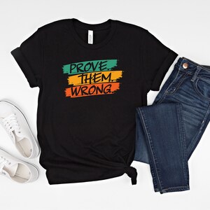 May include: Black t-shirt with the text "PROVE THEM WRONG" in green, orange, and yellow paint strokes.