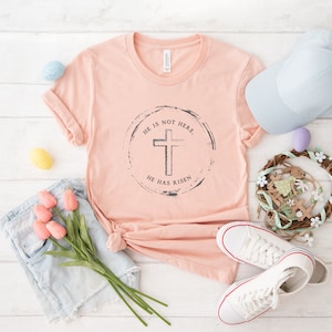 Op de afbeelding: Een perzikkleurig T-shirt met de tekst "HE IS NOT HERE, HE HAS RISEN" rond een kruisafbeelding. Het shirt is gestyled met een spijkerbroek, witte sneakers, een lichtblauwe pet en paasdecoraties, waaronder eieren en een krans.