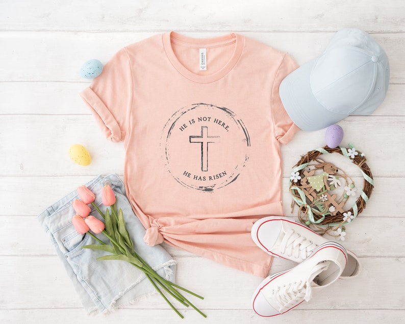 Op de afbeelding: Een perzikkleurig T-shirt met de tekst "HE IS NOT HERE, HE HAS RISEN" rond een kruisafbeelding. Het shirt is gestyled met een spijkerbroek, witte sneakers, een lichtblauwe pet en paasdecoraties, waaronder eieren en een krans.
