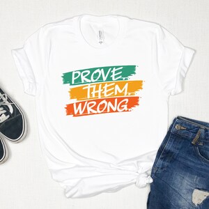 May include: White t-shirt with the text "PROVE THEM WRONG" in white lettering on three colorful stripes. The stripes are green, yellow, and orange.