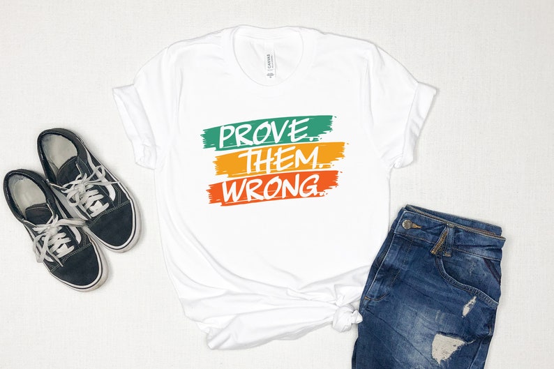 May include: White t-shirt with the text "PROVE THEM WRONG" in white lettering on three colorful stripes. The stripes are green, yellow, and orange.