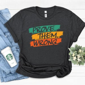 May include: A dark gray t-shirt with the text "PROVE THEM WRONG" in green, yellow, and orange paintbrush strokes.