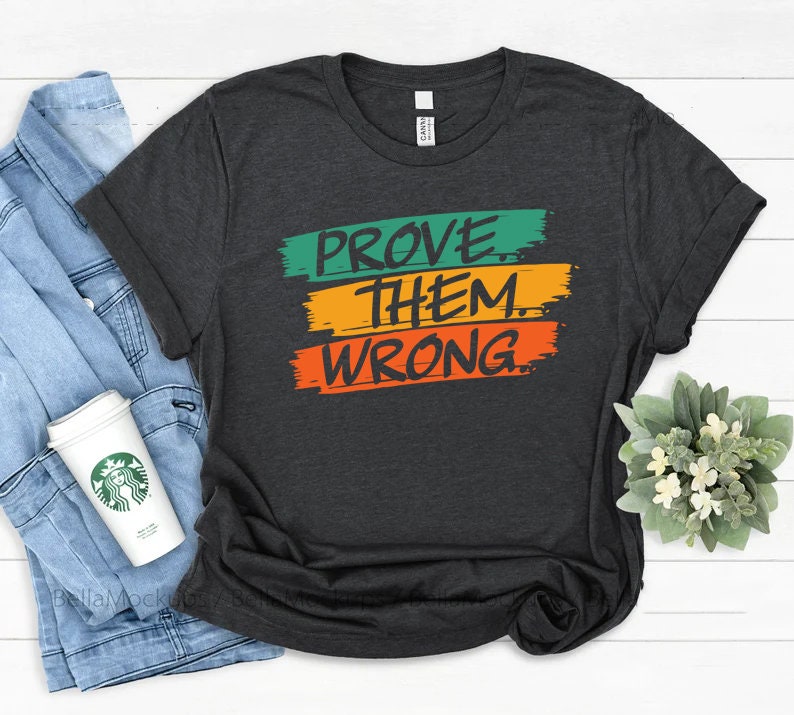 May include: A dark gray t-shirt with the text "PROVE THEM WRONG" in green, yellow, and orange paintbrush strokes.