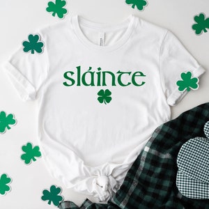 May include: White t-shirt with the Irish word "sl&aacute;inte" printed in green, with a green shamrock underneath. The shirt is surrounded by green shamrock stickers on a white background.
