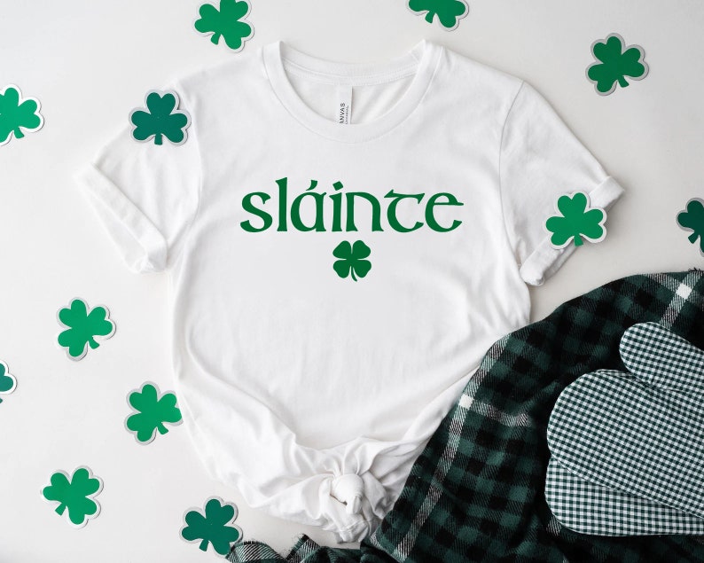 May include: White t-shirt with the Irish word "sl&aacute;inte" printed in green, with a green shamrock underneath. The shirt is surrounded by green shamrock stickers on a white background.