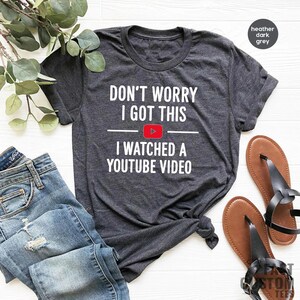 Pode incluir: Uma camiseta cinza escura mesclada com o texto "Don't worry I got this I watched a YouTube video" em branco. Um &iacute;cone de bot&atilde;o de reprodu&ccedil;&atilde;o vermelho est&aacute; entre as palavras "this" e "I".