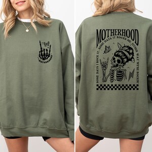 May include: Green sweatshirt with a black graphic print of a skeleton with a butterfly and the text "MOTHERHOOD. SOME DAYS I ROCK IT. SOME DAYS IT ROCKS ME. EITHER WAY WE'RE ROCKIN'" on the back. The front of the sweatshirt has a smaller graphic print of a skeleton hand with a butterfly and the text "MOTHERHOOD".