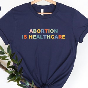 K&ouml;nnte beinhalten: Ein marineblaues T-Shirt mit dem Aufdruck "ABORTION IS HEALTHCARE" in Regenbogenfarben.