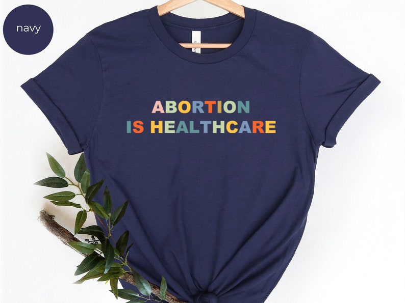 K&ouml;nnte beinhalten: Ein marineblaues T-Shirt mit dem Aufdruck "ABORTION IS HEALTHCARE" in Regenbogenfarben.