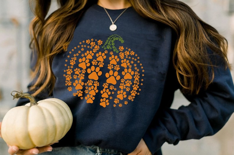 May include: A navy blue sweatshirt with a pumpkin design made of orange paw prints. The pumpkin has a green stem and leaves.