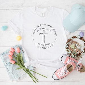 Op de afbeelding: Wit T-shirt met een zwart kruisontwerp in een cirkel, met de tekst "HE IS NOT HERE. HE HAS RISEN." De afbeelding bevat ook een lichtblauwe baseballpet, roze sneakers en een kleine krans met paasversieringen.