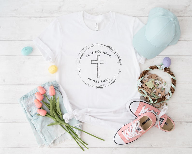 Op de afbeelding: Wit T-shirt met een zwart kruisontwerp in een cirkel, met de tekst "HE IS NOT HERE. HE HAS RISEN." De afbeelding bevat ook een lichtblauwe baseballpet, roze sneakers en een kleine krans met paasversieringen.