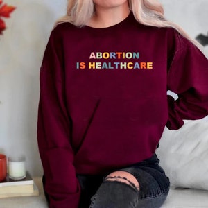 K&ouml;nnte beinhalten: Ein weinroter Pullover mit dem Text "ABORTION IS HEALTHCARE" in Regenbogenfarben.