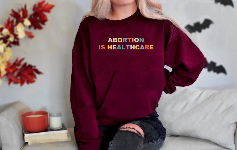K&ouml;nnte beinhalten: Ein weinroter Pullover mit dem Text "ABORTION IS HEALTHCARE" in Regenbogenfarben.