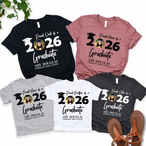 May include: Assorted t-shirts with graduation-themed designs. The shirts are black, pink, gray, white, and dark gray. Each shirt features the text "Proud Dad of a 2026 Graduate", "Proud Mom of a 2026 Graduate", "Proud Niece of a 2026 Graduate", "Proud Brother of a 2026 Graduate", and "Proud Sister of a 2026 Graduate".