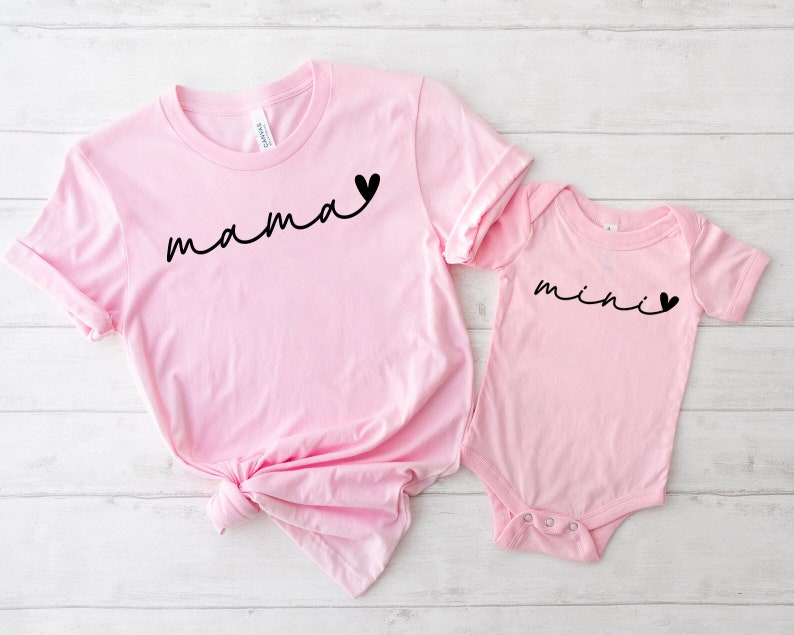 May include: Pink t-shirt with the word "mama" in black script with a small black heart. A pink baby onesie with the word "mini" in black script with a small black heart.