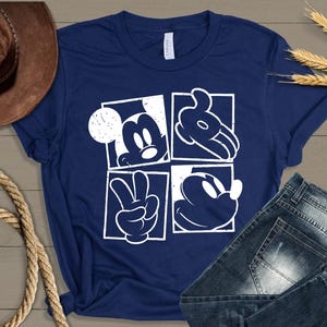 May include: Navy blue t-shirt featuring a white graphic of Mickey Mouse in four square panels. The design includes Mickey's face, hands, and silhouette. The shirt is paired with denim jeans and a brown cowboy hat.