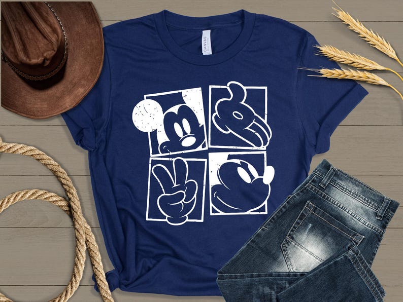 May include: Navy blue t-shirt featuring a white graphic of Mickey Mouse in four square panels. The design includes Mickey's face, hands, and silhouette. The shirt is paired with denim jeans and a brown cowboy hat.
