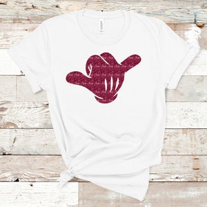 May include: White t-shirt with a burgundy glitter hand making the shaka sign.