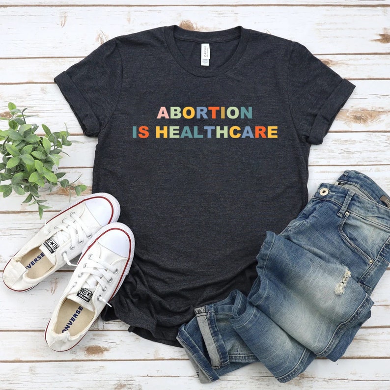 K&ouml;nnte beinhalten: Ein dunkelgraues T-Shirt mit dem Text "ABORTION IS HEALTHCARE" in Regenbogenfarben. Das Shirt liegt flach auf einer Holzoberfl&auml;che mit einem Paar wei&szlig;en Turnschuhen und einer blauen Jeans.