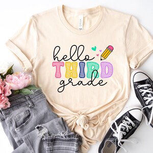 Pode incluir: Uma camiseta bege com o texto "hello third grade" em letras coloridas e um desenho de lápis. A camiseta está colocada sobre uma superfície branca com um par de tênis pretos e um par de calças jeans.