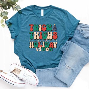 Puede incluir: Una camiseta azul turquesa con el texto "THICK THIGHS and HOLIDAY VIBES" en una fuente retro con colores rojo, verde y blanco. El texto est&aacute; centrado en la camiseta.