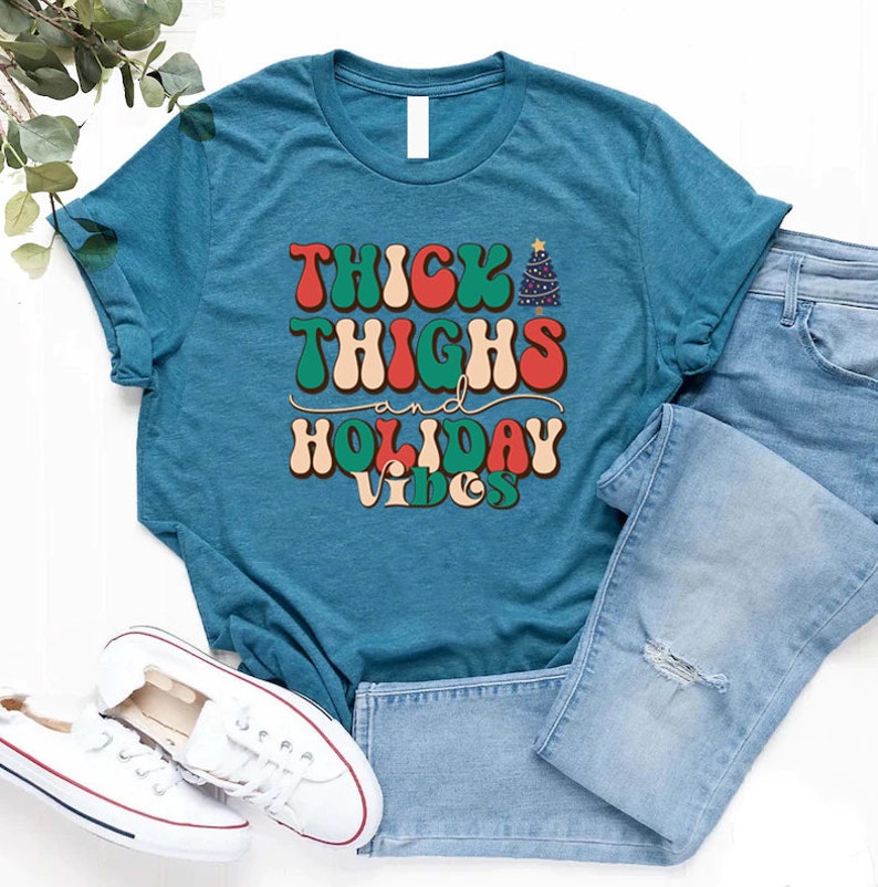 Puede incluir: Una camiseta azul turquesa con el texto "THICK THIGHS and HOLIDAY VIBES" en una fuente retro con colores rojo, verde y blanco. El texto est&aacute; centrado en la camiseta.