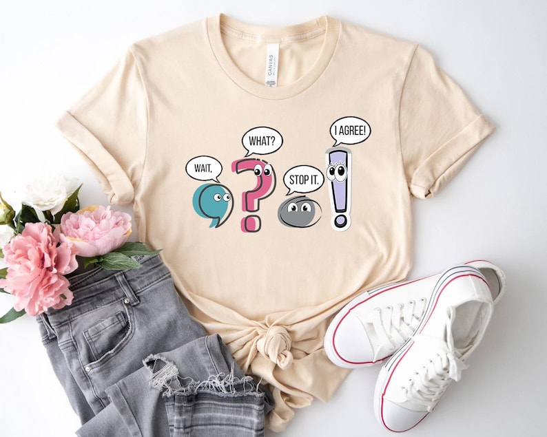 May include: A beige t-shirt with a graphic of punctuation marks with faces and speech bubbles. The speech bubbles say "Wait", "What?", "Stop it", and "I agree!"