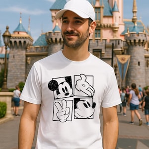 May include: White t-shirt featuring a four-panel design with Mickey Mouse imagery. The panels include Mickey's face, hands, and silhouette. The person in the image is wearing a white baseball cap. The background shows a castle.
