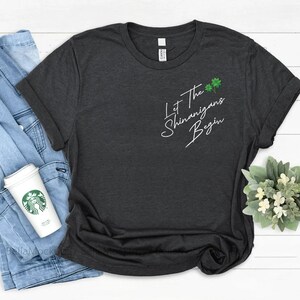 Peut inclure: Un t-shirt gris fonc&eacute; chin&eacute; avec le texte "Let the Shenanigans Begin" en &eacute;criture blanche. Le texte est centr&eacute; sur le t-shirt et est accompagn&eacute; d'un petit graphique de tr&egrave;fle vert.