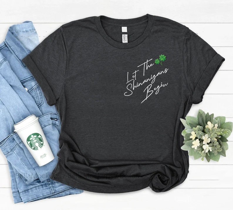 Peut inclure: Un t-shirt gris fonc&eacute; chin&eacute; avec le texte "Let the Shenanigans Begin" en &eacute;criture blanche. Le texte est centr&eacute; sur le t-shirt et est accompagn&eacute; d'un petit graphique de tr&egrave;fle vert.