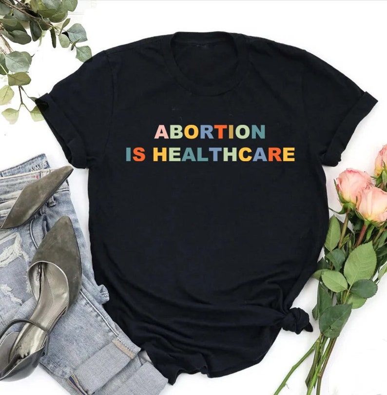 K&ouml;nnte beinhalten: Schwarzes T-Shirt mit dem Text "ABORTION IS HEALTHCARE" in Regenbogenfarben.